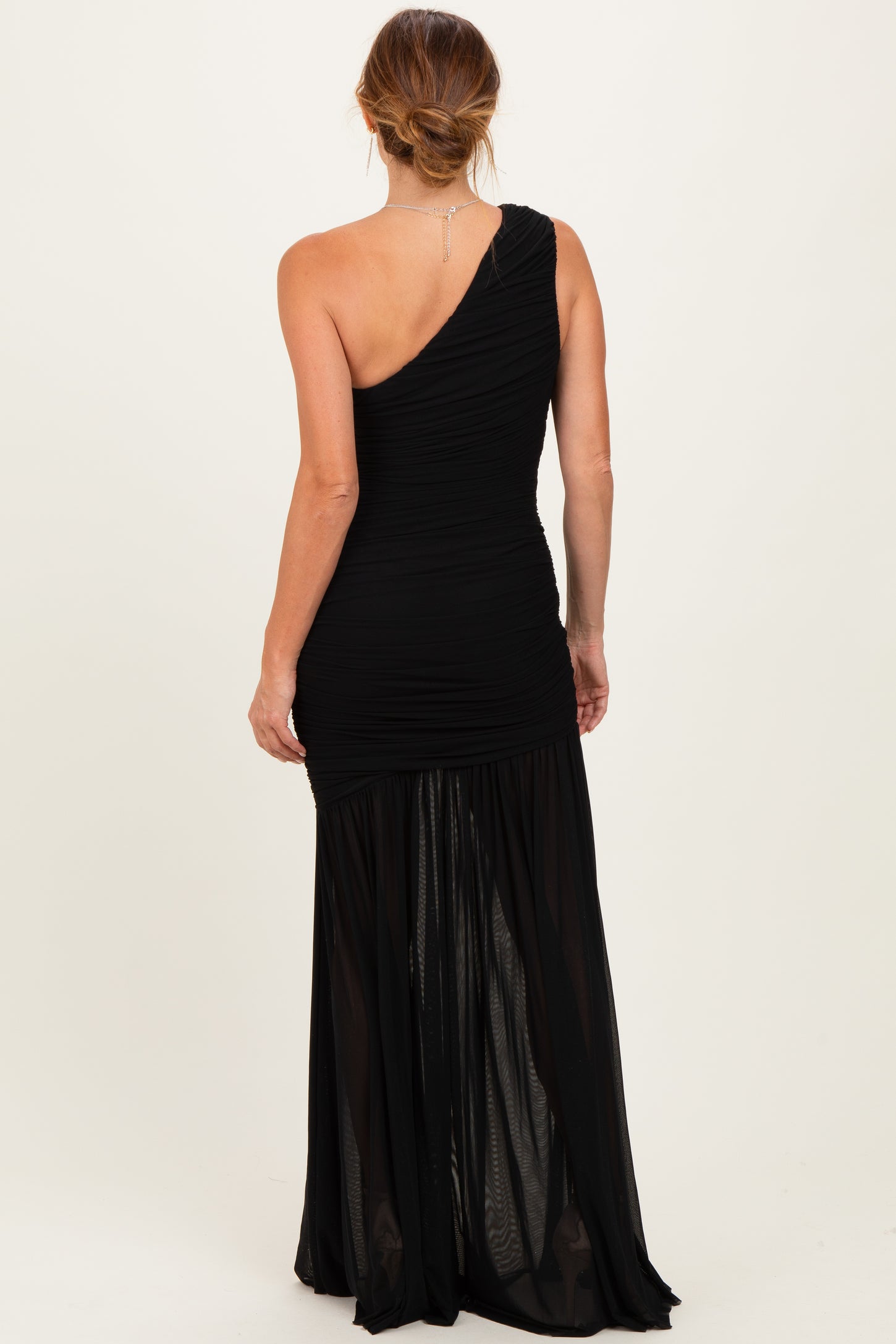 woman wearing a black one-shoulder evening gown with ruched detailing and a flowing sheer skirt from the back