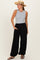 woman wearing a gray sleeveless top and black wide-leg pants, accessorized with a straw hat, standing with one hand in her pocket