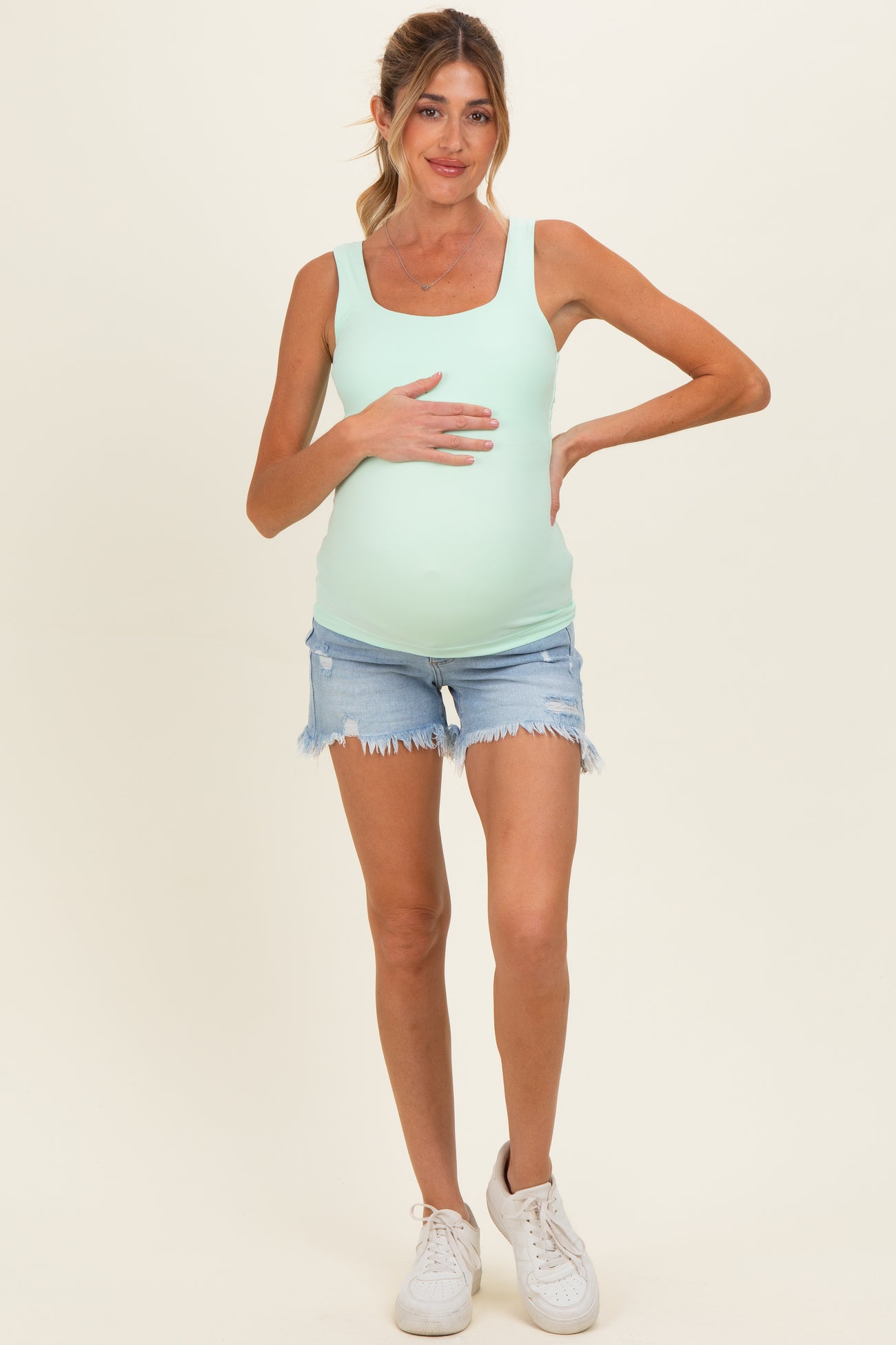 woman wearing a light green maternity tank top and distressed denim shorts, holding her baby bump