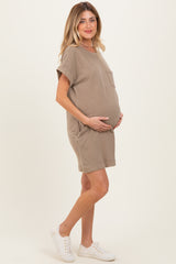 woman wearing a taupe short sleeve maternity dress, gently cradling her baby bump while standing against a light background
