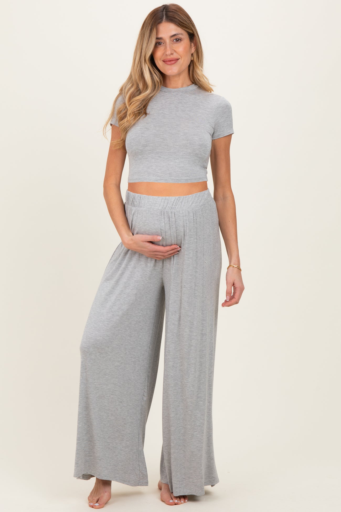woman wearing a gray maternity crop top and matching wide-leg pants, gently holding her baby bump
