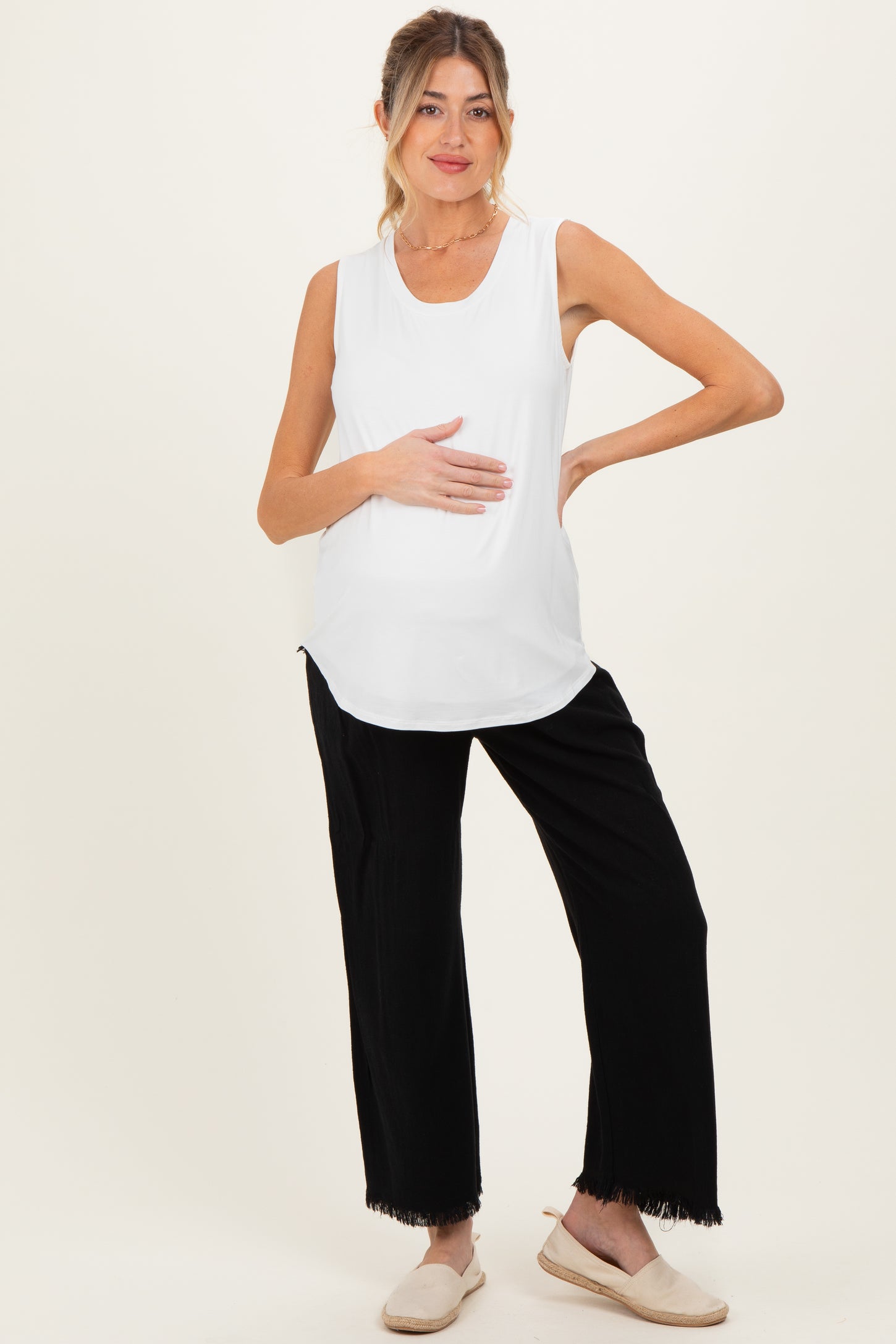 woman wearing a white sleeveless maternity top and black fringe hem pants, holding her baby bump