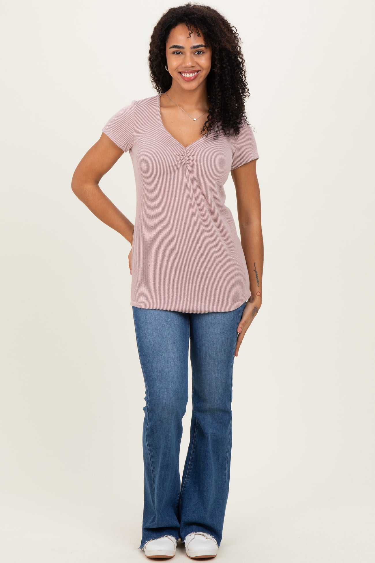 woman wearing a light pink ribbed short sleeve top and blue flared jeans, standing and smiling
