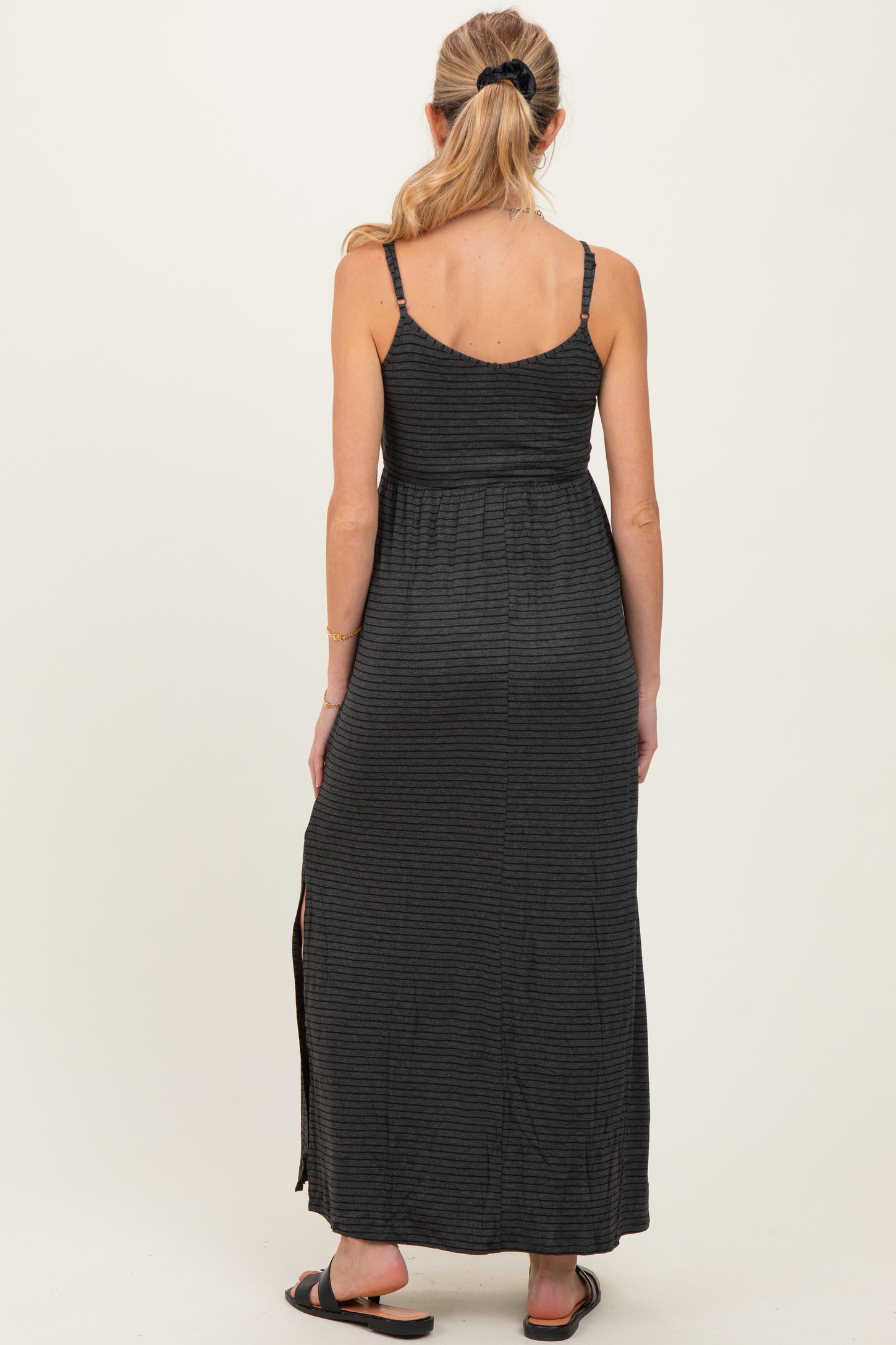 woman wearing a black striped maternity maxi dress with adjustable straps and a side slit, viewed from the back