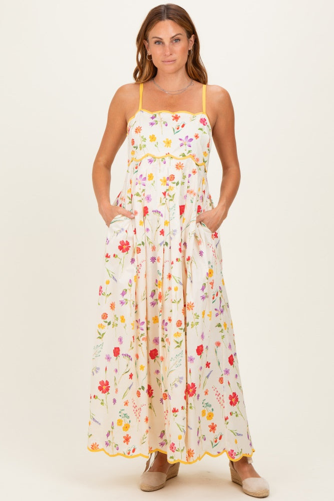 woman wearing a white floral maxi dress with adjustable straps and pockets, styled with scalloped hem details