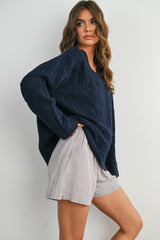 woman wearing a dark blue cable knit oversized sweater and striped shorts, posing against a white background