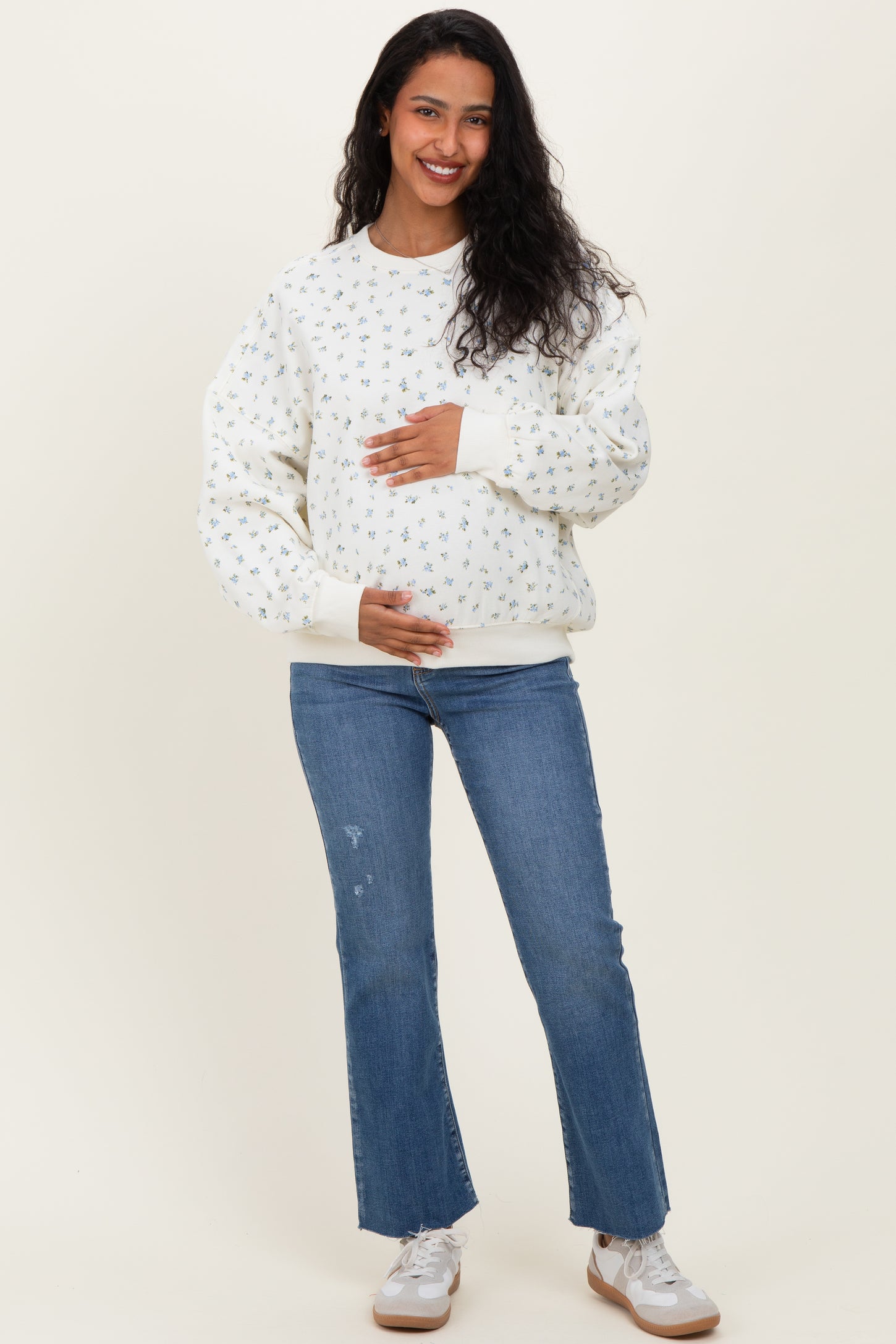 woman wearing a white floral print maternity sweatshirt and blue jeans, holding her baby bump