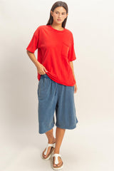 woman wearing a red oversized t-shirt with pocket and blue wide-leg shorts, standing in casual footwear