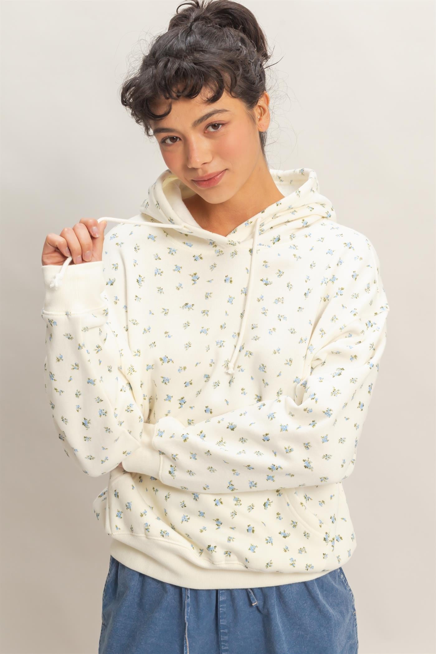 woman wearing a cream floral hoodie with a drawstring and long sleeves, posing with one hand on her shoulder