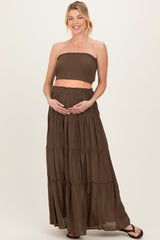 woman wearing a brown maternity two-piece outfit with a smocked strapless top and a flowy maxi skirt, holding her baby bump