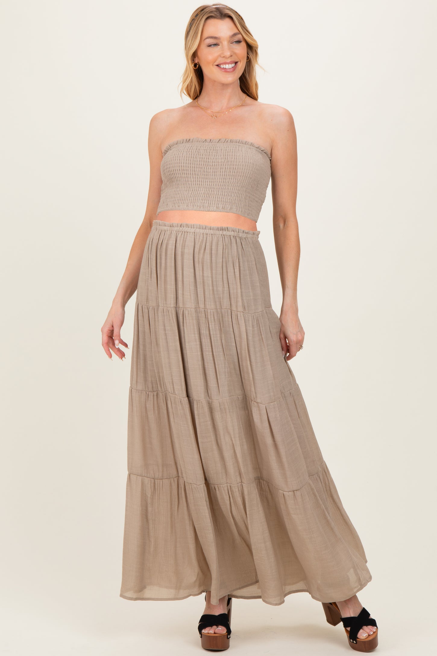 woman wearing a beige strapless smocked maternity crop top and matching maxi skirt, smiling with hands on hips