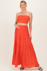 woman wearing a bright orange maternity two-piece set with a smocked bandeau top and a tiered maxi skirt, holding her baby bump