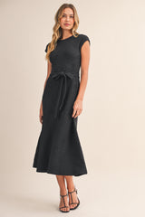 woman wearing a black cap sleeve midi dress with a floral texture and a tie belt, standing in a neutral background