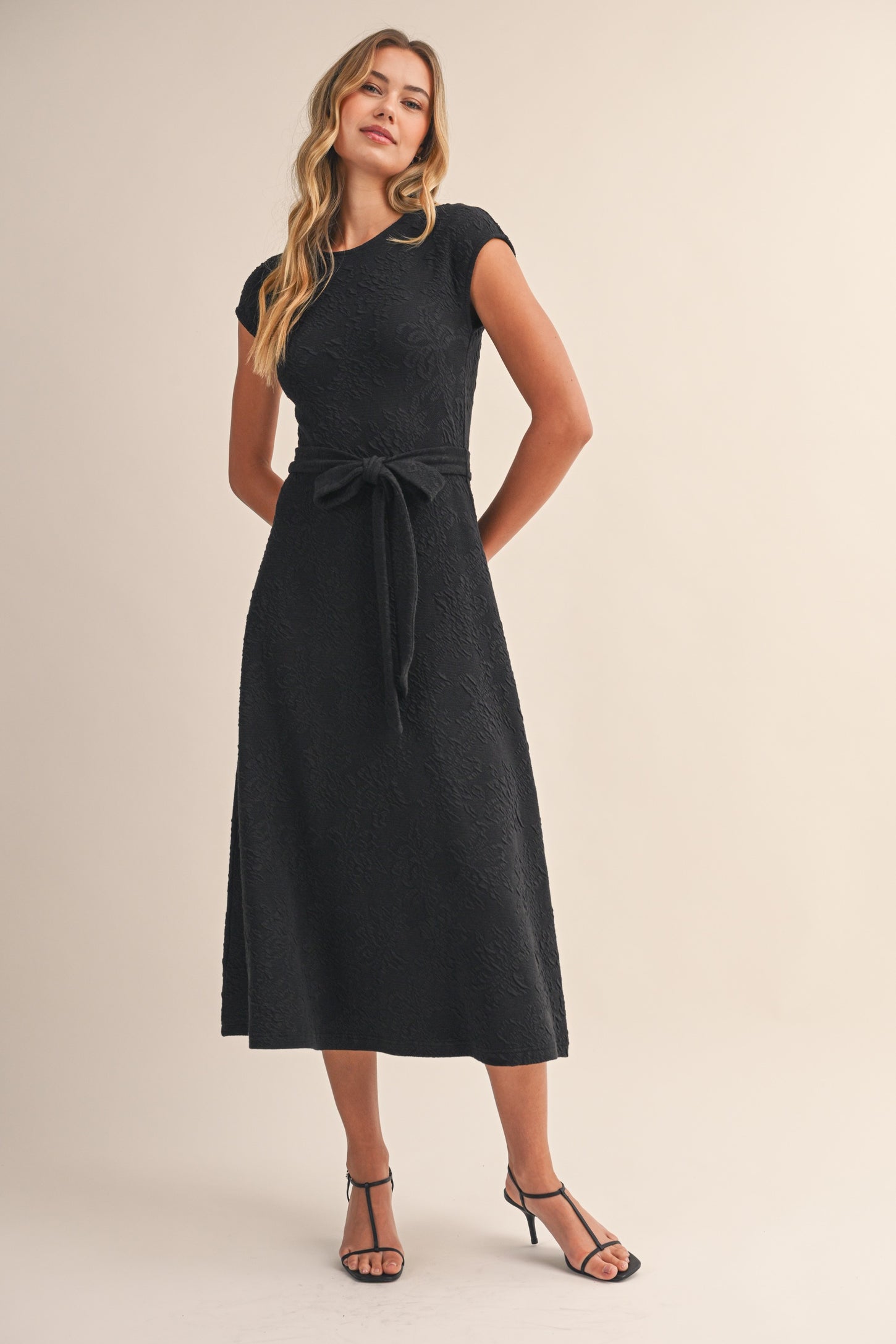 woman wearing a black mid-length dress with a fitted bodice and a tie belt, styled with strappy sandals