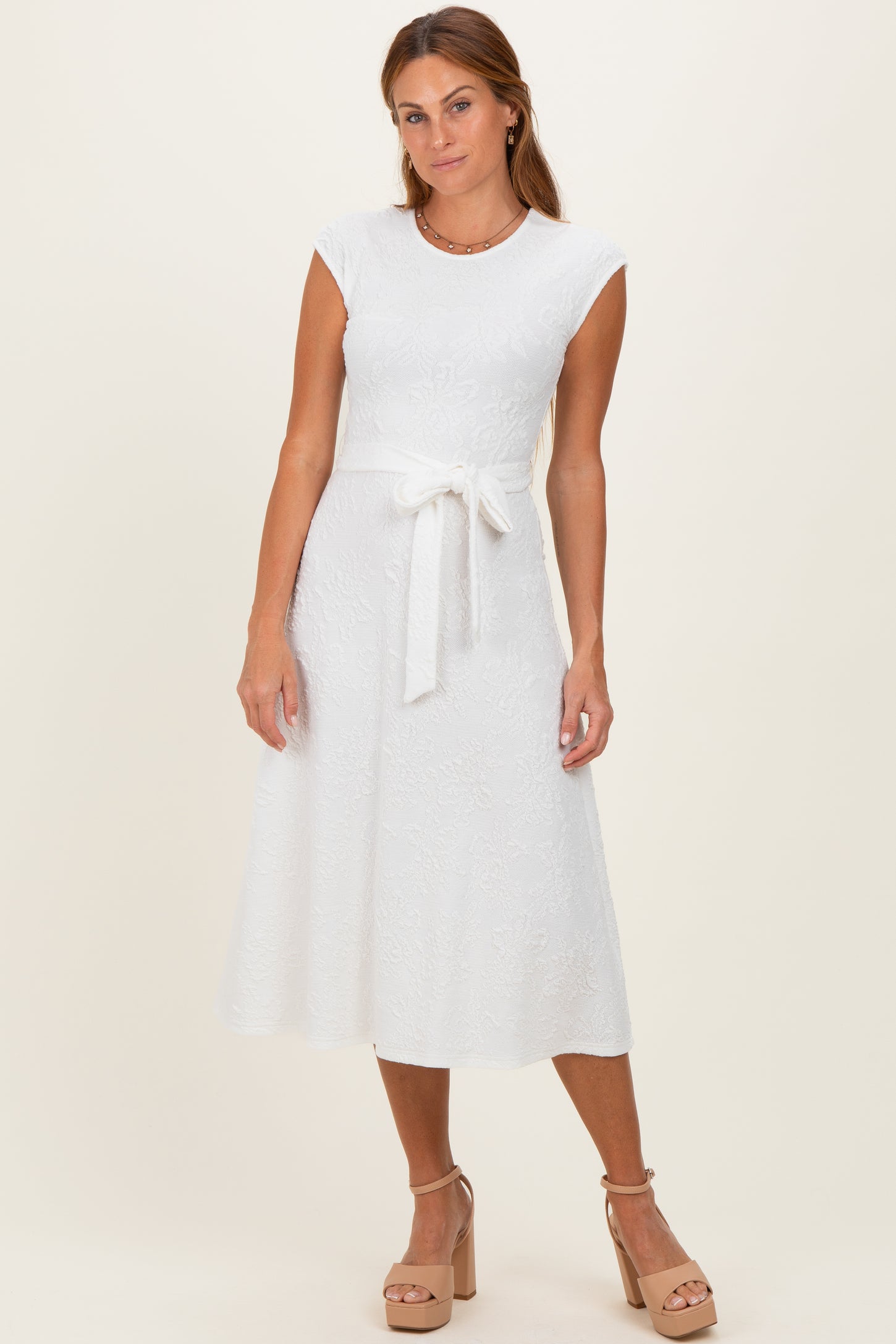 woman wearing a white cap sleeve midi dress with a tie waist and textured floral pattern, posing with a neutral backdrop