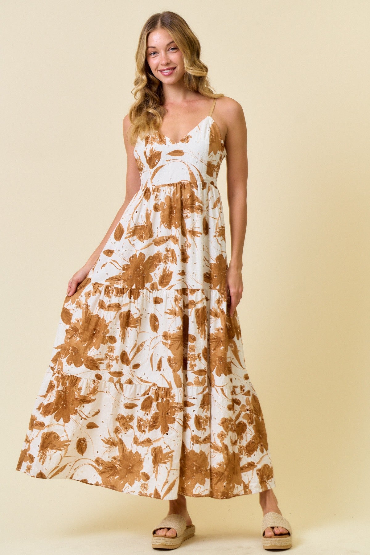 woman wearing a white maxi dress with brown floral prints and spaghetti straps, standing against a light beige background