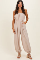 woman wearing a beige strapless jumpsuit with a drawstring waist and side pockets, styled with heeled sandals