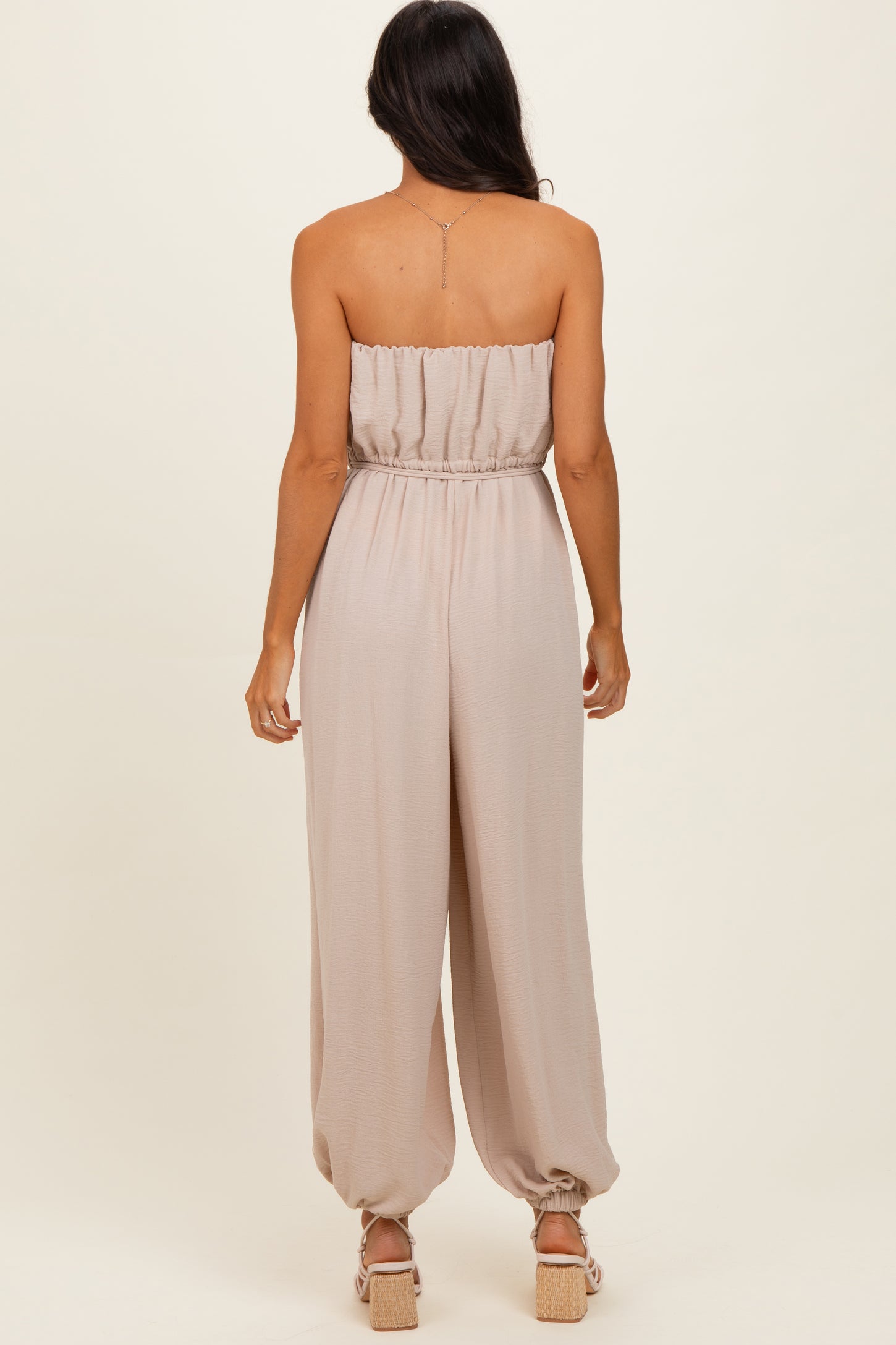 woman wearing a strapless beige jumpsuit with a gathered waistband and relaxed fit, showing the back view