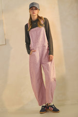 woman wearing pink and white striped overalls with a dark long sleeve top and a gray cap, styled with casual sneakers