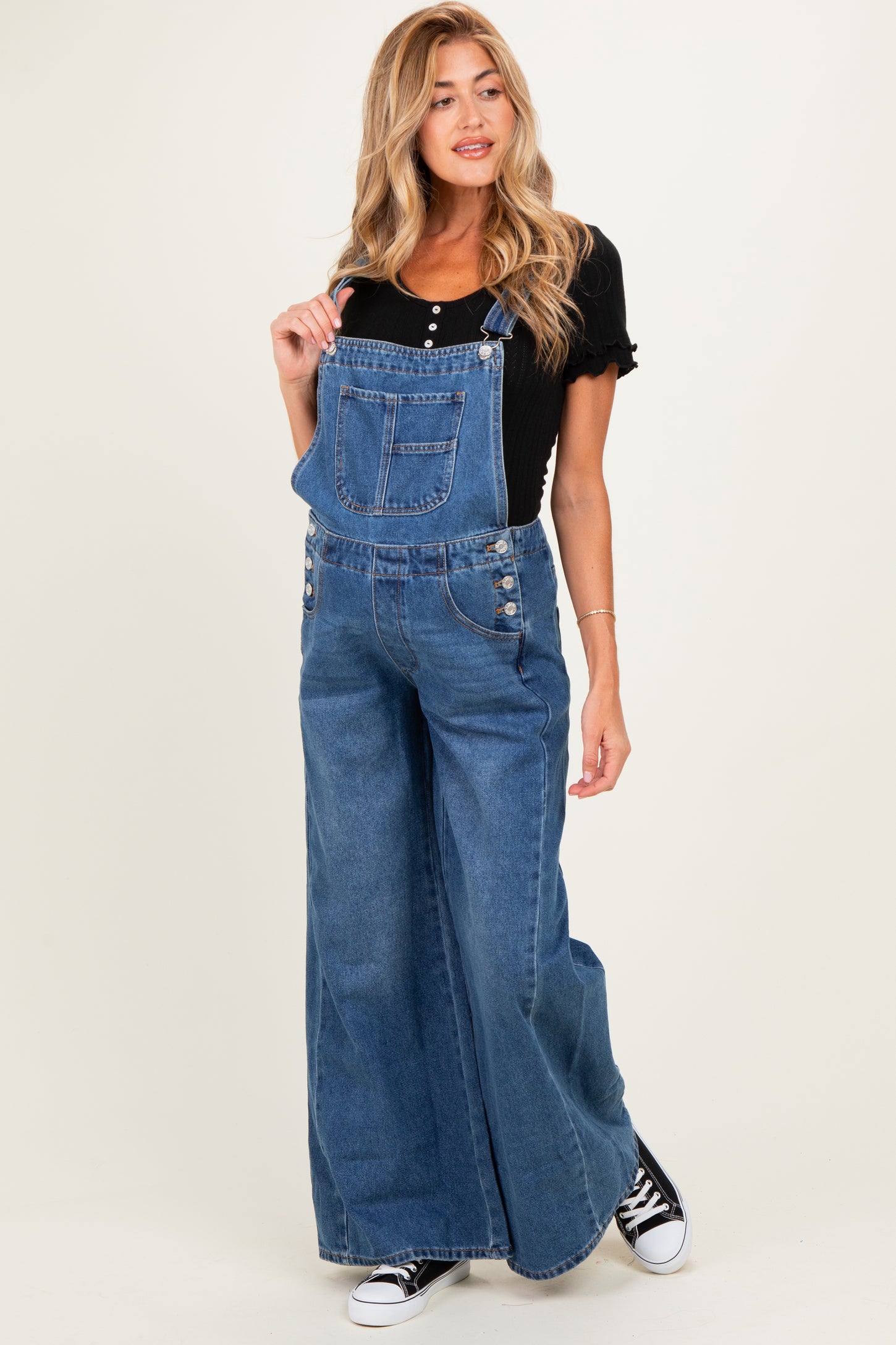 woman wearing a black short sleeve maternity top and denim overalls, posing with one shoulder strap down