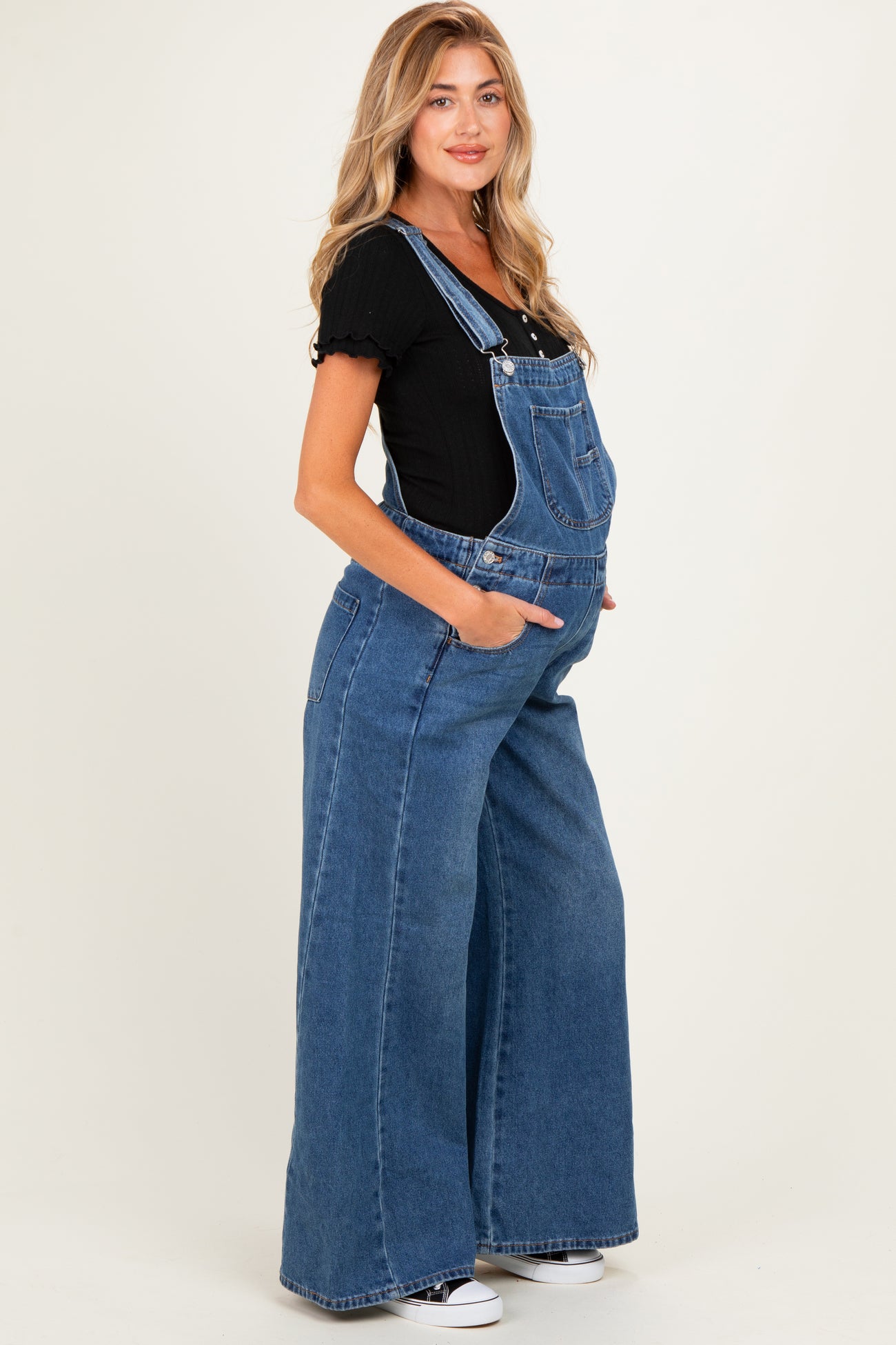 woman wearing a black short sleeve top and blue wide leg maternity overalls, showing her baby bump