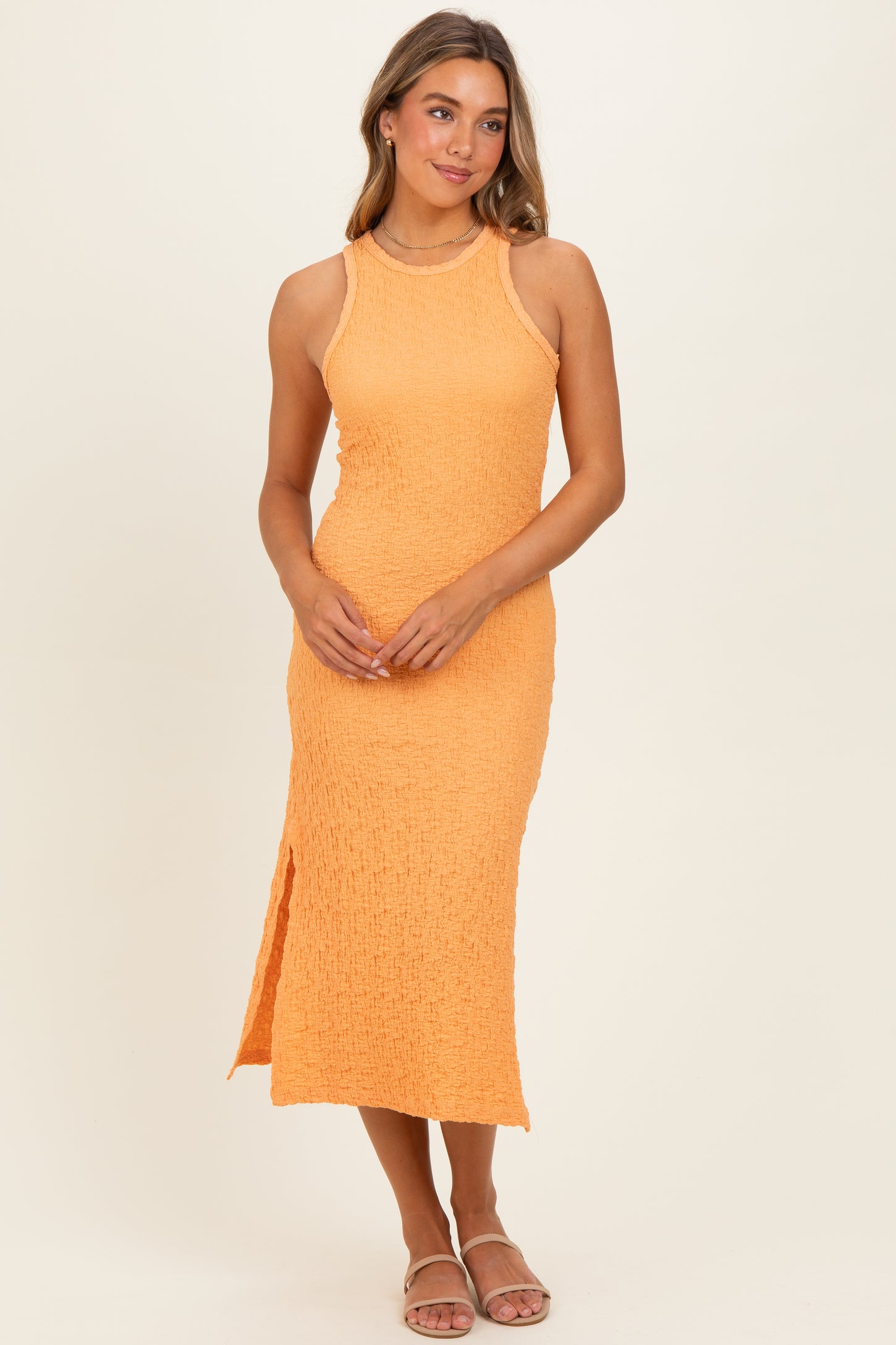 woman wearing a sleeveless orange midi dress with a fitted bodice and side slit, standing against a neutral background