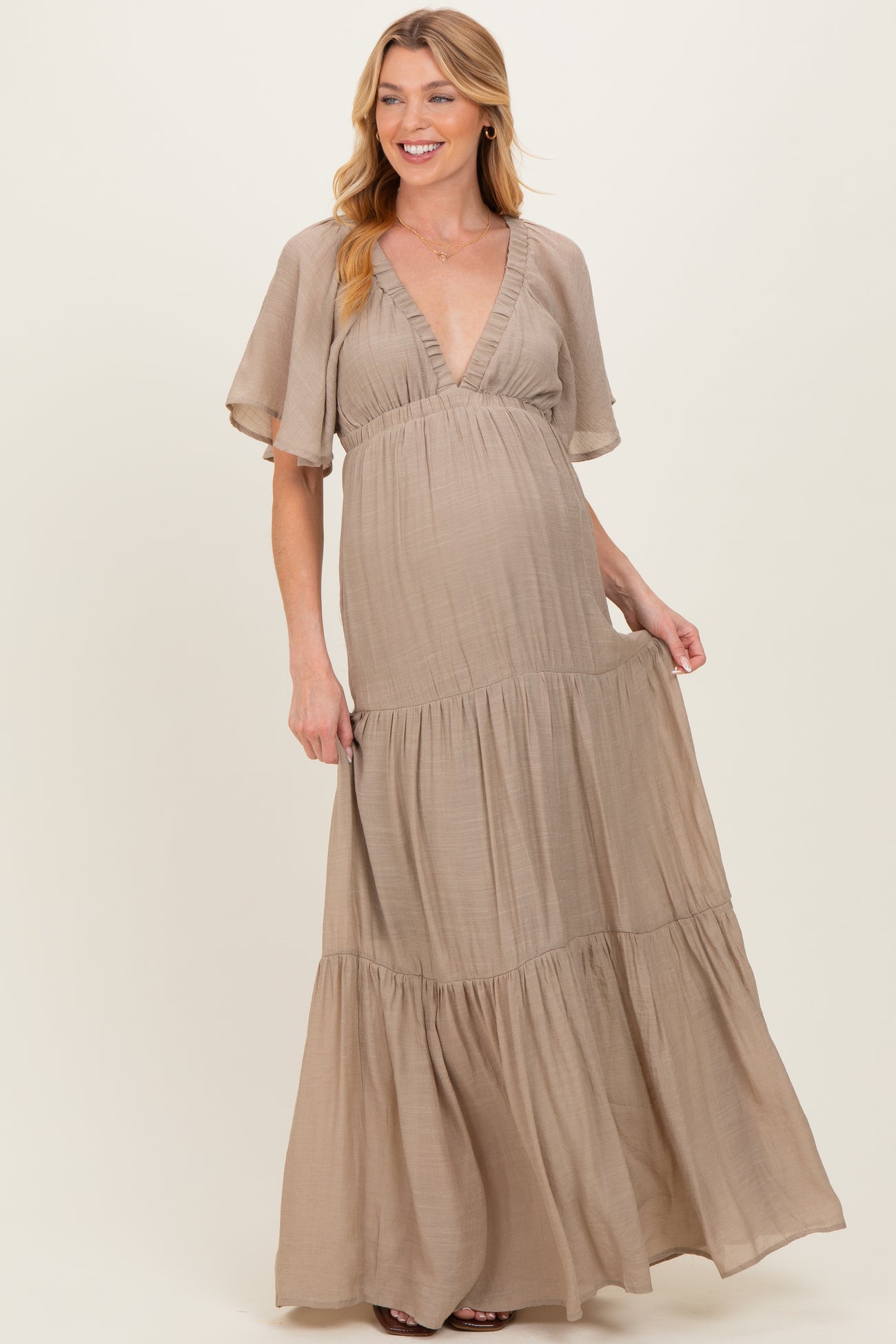 woman wearing a beige v-neck maxi maternity dress with flutter sleeves and tiered skirt, showing her baby bump