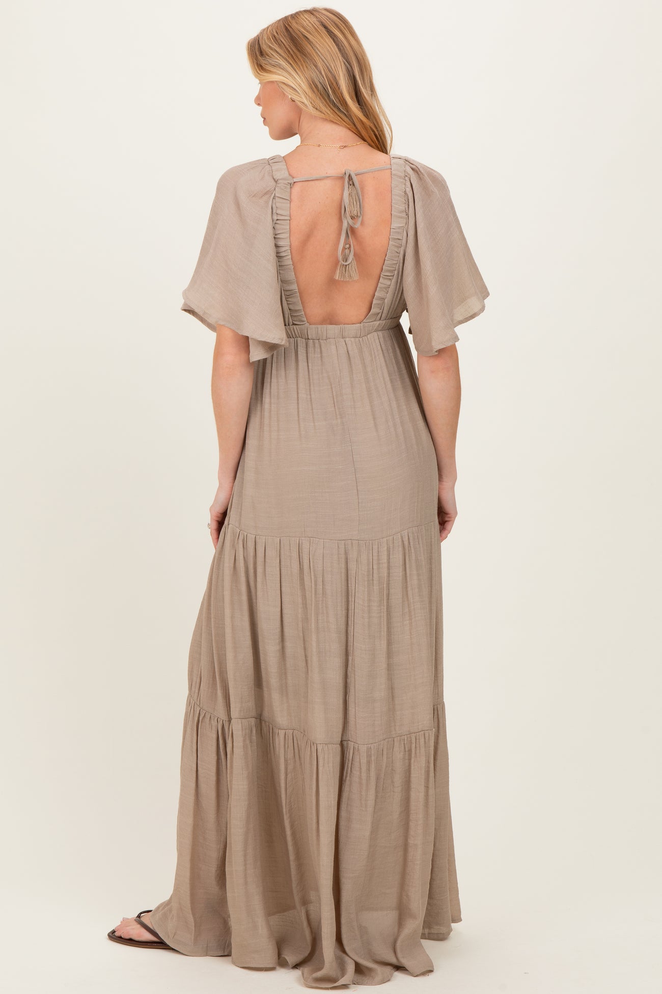 woman wearing a beige maternity maxi dress with short flutter sleeves and open back design