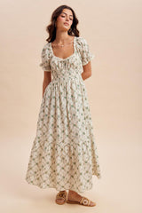 woman wearing a floral print maxi dress with short puff sleeves and a ruched bodice, standing against a neutral background