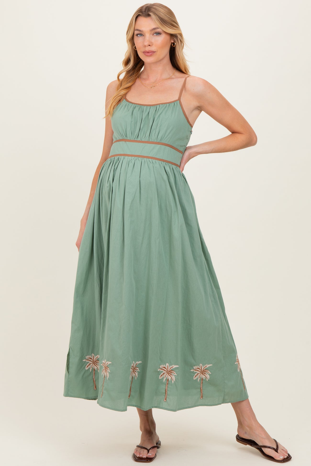 woman wearing a green maternity maxi dress with a cinched waist and palm tree embroidery at the hem