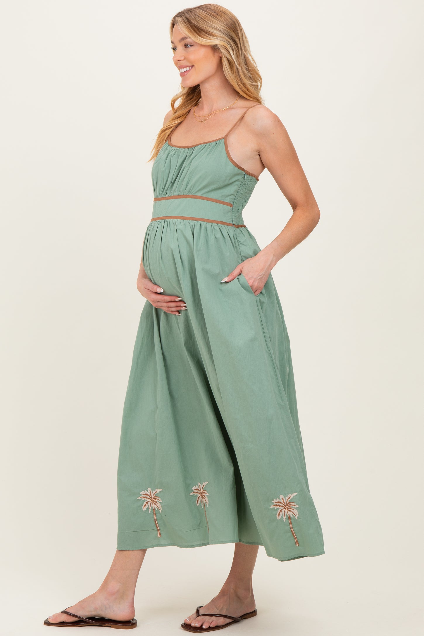 woman wearing a green maternity maxi dress with palm tree embroidery and pockets, cradling her baby bump