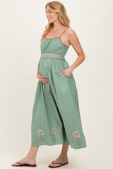 woman wearing a green maternity maxi dress with palm tree embroidery and pockets, cradling her baby bump