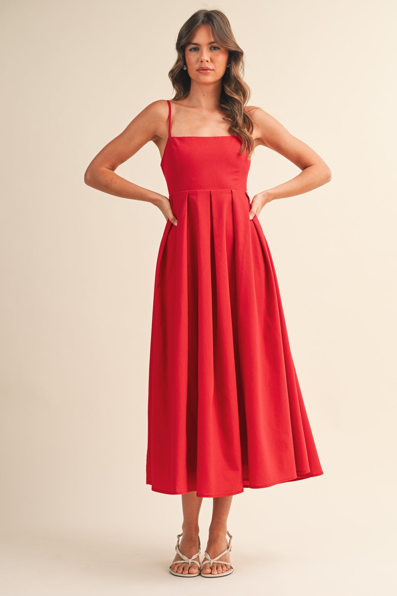 woman wearing a red spaghetti strap midi dress with a pleated skirt and posing with hands on hips