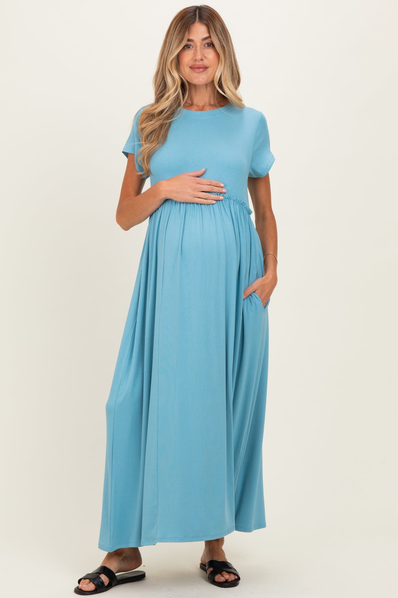 woman wearing a light blue full-length maternity dress with short sleeves and side pockets, holding her baby bump