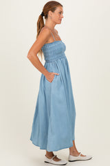 woman wearing a light blue maxi dress with smocked bodice and pockets, standing sideways with long hair tied back