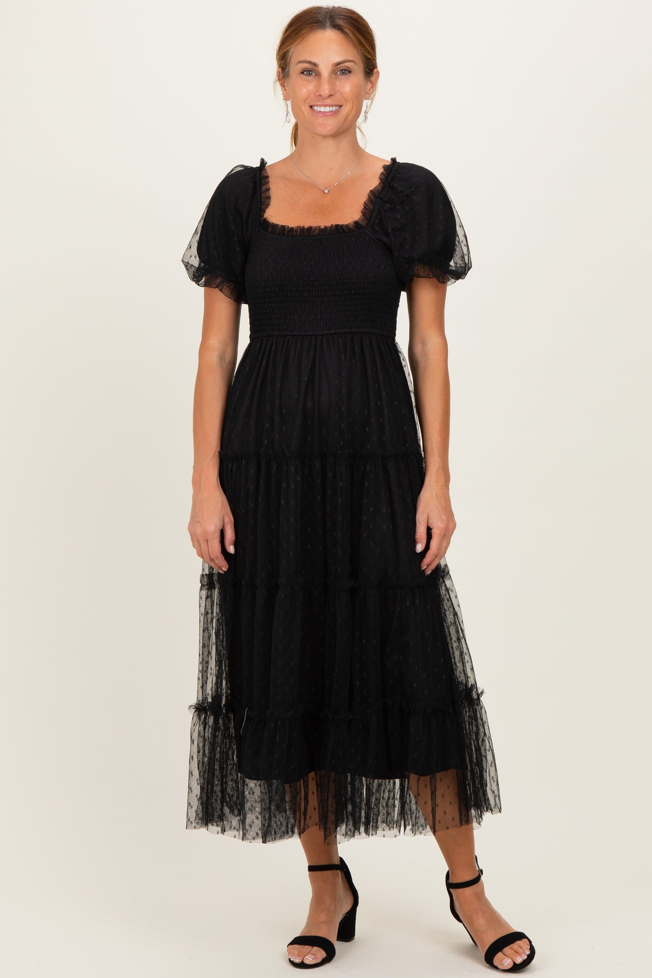 woman wearing a black tiered puff sleeve maxi dress with a smocked bodice and sheer overlay, standing in black heels
