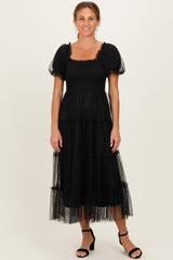 woman wearing a black tiered puff sleeve maxi dress with a smocked bodice and sheer overlay, standing in black heels