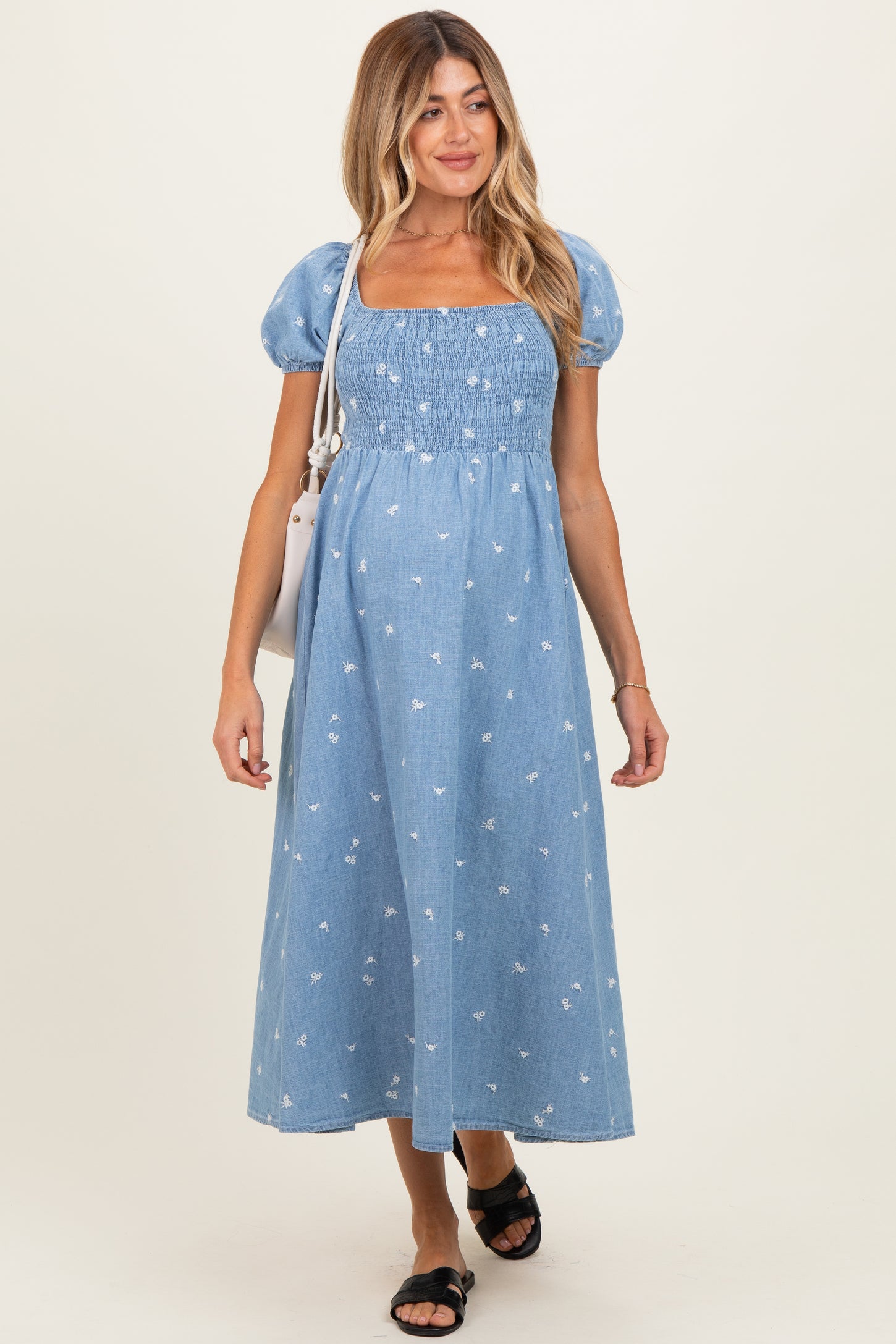 woman wearing a light blue maternity dress with puff sleeves and floral patterns, smiling while carrying a bag