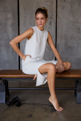 woman wearing a light gray sleeveless midi dress with side slits, seated on a bench in a casual pose