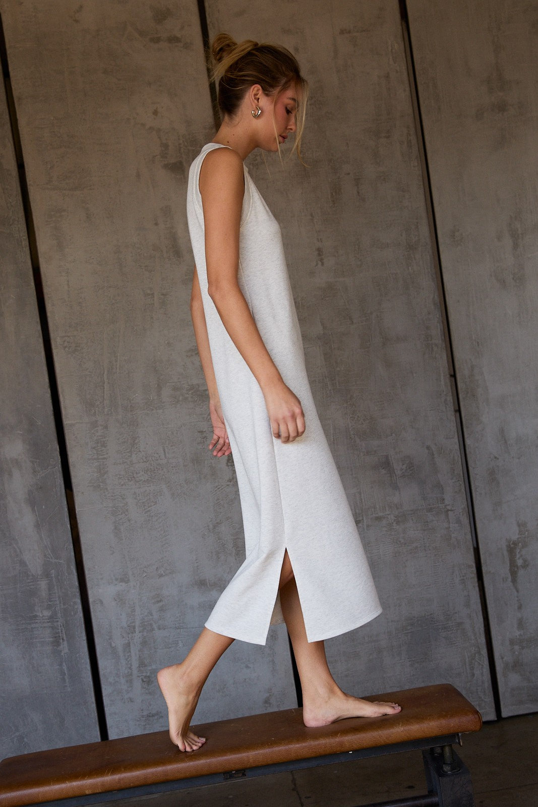 woman wearing a light gray sleeveless maxi dress with side slits, standing barefoot on a wooden bench