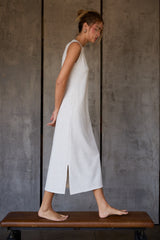 woman wearing a light gray sleeveless maxi dress with side slits, standing barefoot on a bench