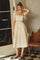 woman wearing a cream-colored midi dress with ribbon straps and scalloped detailing, styled with a clutch purse