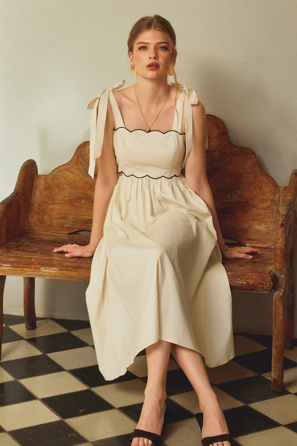 woman wearing a cream midi dress with tie straps and black trim, sitting on a wooden bench