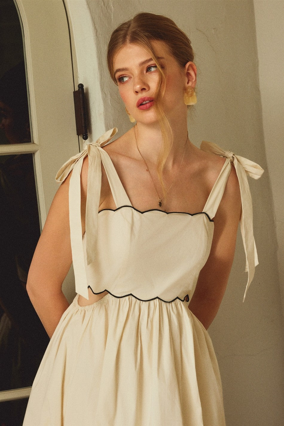 woman wearing a cream square neck dress with scalloped edges and tied straps, styled with subtle makeup and earrings