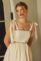 woman wearing a cream square neck dress with scalloped edges and tied straps, styled with subtle makeup and earrings