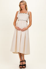 woman wearing a cream maternity dress with scalloped straps, holding her baby bump and standing in a light studio