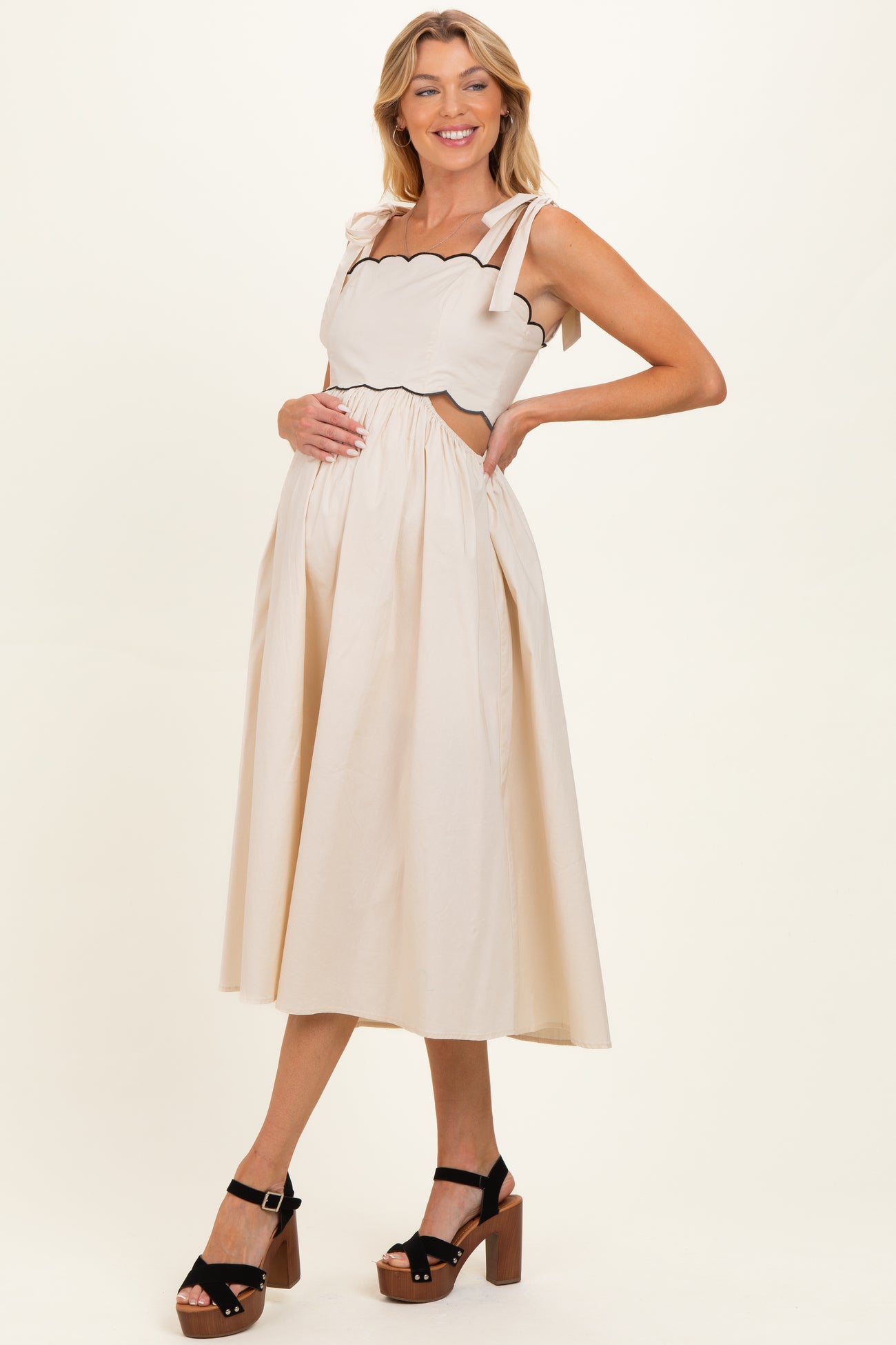 woman wearing a beige maternity dress with scalloped edges and tie straps, showcasing her baby bump, paired with black platform sandals