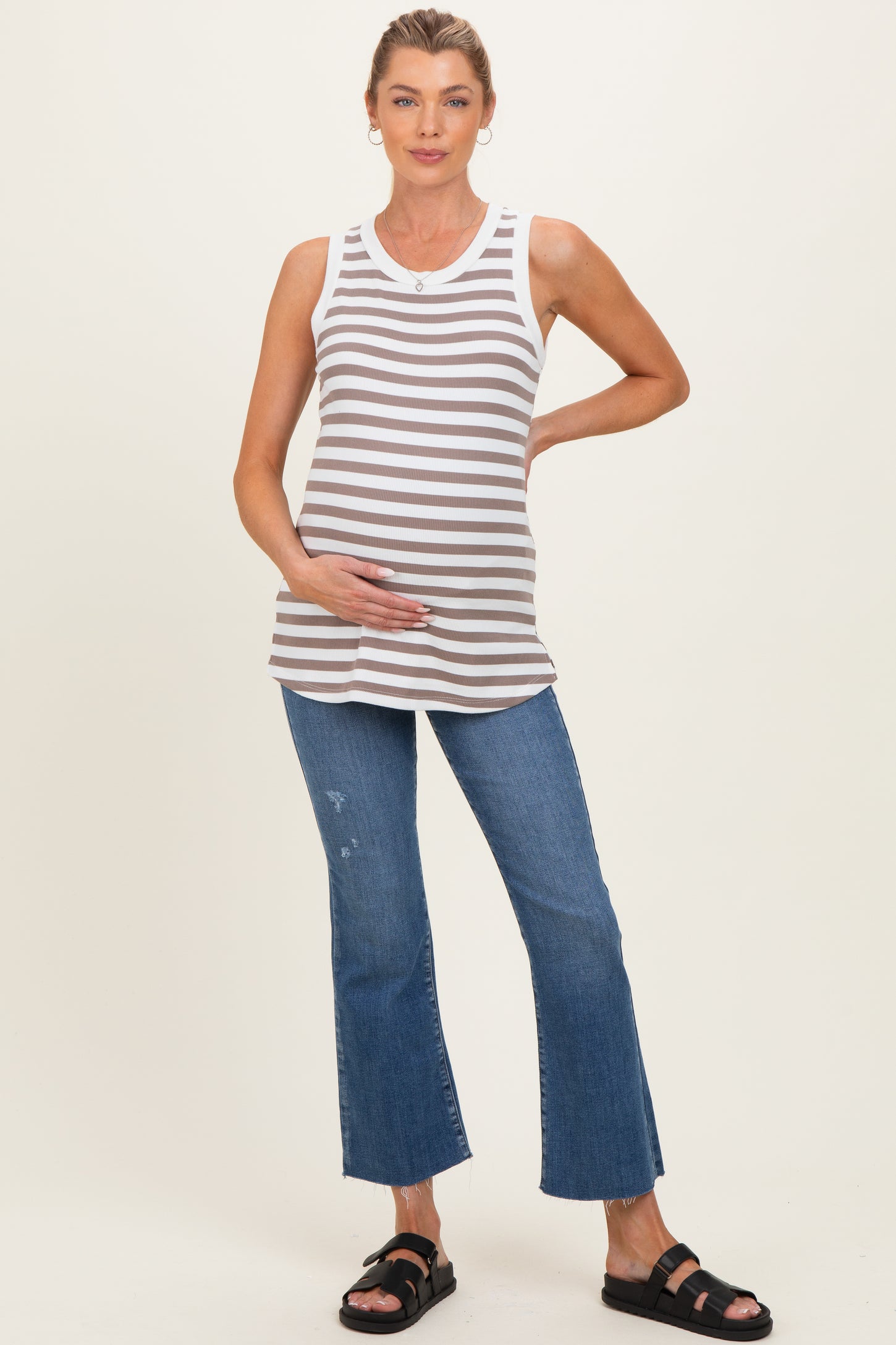 woman wearing a gray and white striped maternity tank top and distressed blue flared jeans, gently holding her baby bump