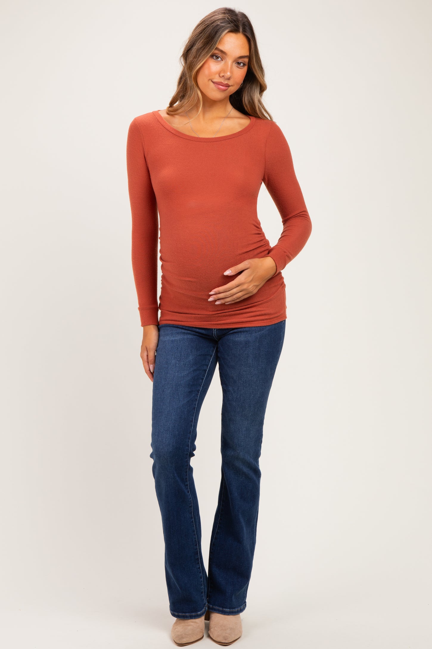 woman wearing a rust long sleeve maternity top and blue flared jeans, gently touching her baby bump