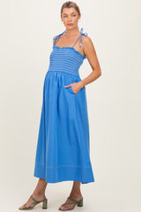 woman wearing a blue maternity maxi dress with smocked bodice and tie straps, featuring pockets and a flowing skirt